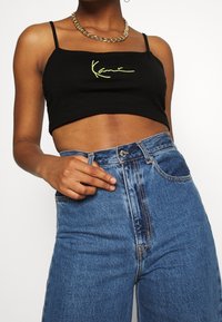 Svart kort linne med gul broderad text, kombinerat med höga blå jeans i denim med dragkedja och knappstängning.