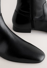 Schwarze Leder-Stiefeletten mit Reißverschluss an der Seite und niedrigem Absatz, gezeigt vor hellem neutralem Hintergrund.