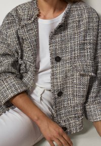 Beżowa i niebieska marynarka w stylu tweed, charakteryzująca się prostokątnym krojem, postrzępionymi brzegami i ciemnymi guzikami. Noszona na białej koszulce.