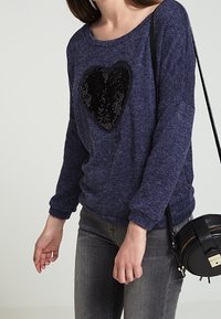 Mörkblå stickad tröja med ett svart paljettformad hjärta i mitten, ihopparad med grå jeans och en svart axelremsväska.