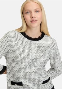 Weiß und schwarz gewebter Pullover mit strukturierter Oberfläche, ausgestattet mit einem fransigen schwarzen Kragen und zwei Fronttaschen mit schwarzen Akzenten.