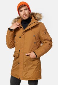 Parka matelassée marron avec capuche doublée de fausse fourrure, devant boutonné, plusieurs poches et accents en métal noir. Portée sur un turtleneck noir.