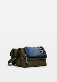 Borsa trapuntata verde oliva con pattina in denim, dotata di zip e patch con logo. Design compatto con tracolla regolabile e dettagli testurizzati.