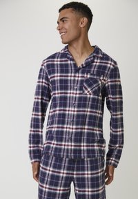 Joven sonriendo, vistiendo pijamas de cuadros azul, blanco y rojo con botones y un bolsillo en el pecho, de pie contra un fondo claro y liso.