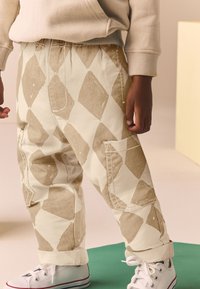 Beiget, timanttikuviolliset löysät housut, joissa on joustava vyötärö, taskut ja käännetyt lahkeet; yhdistetty valkoisiin lenkkareihin. Sileä kangaspinta.