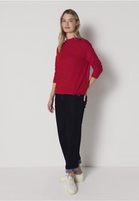Roter Pullover mit Rundhalsausschnitt und langen Ärmeln, kombiniert mit schwarzen, gekrempelten Hosen. Die Schuhe sind weiße Sneaker mit einer hellen Sohle.