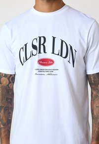 Bijela pamučna majica s podebljanim crnim natpisom "CLSR LDN" i crvenim ovalnim logotipom "Closure Ldn." Okrugli izrez, kratki rukavi, opušteni kroj.