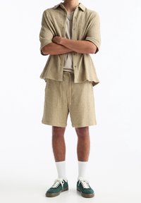 Beiges, strukturiertes Kurzarmhemd und passende Shorts, kombiniert mit einem hellen T-Shirt, weißen Socken und grünen Turnschuhen.