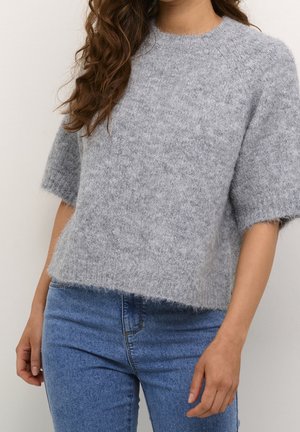 Frau trägt einen flauschigen grauen Kurzarmpullover und blaue hochgeschnittene Jeans, mit lockigem, welligem braunem Haar über einer Schulter.