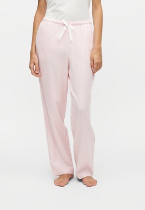 Femme portant un pantalon de pyjama ample rose clair avec un cordon blanc, debout pieds nus sur un fond blanc uni.