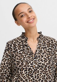 Leopardmönstrad skjorta med texturerad tyg, med en svart och beige färgschema, en volangkrage och en V-ringning design.