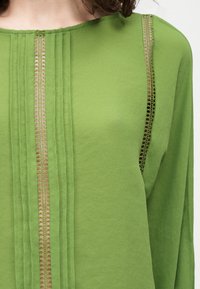 Πράσινο μακρυμάνικο μπλουζάκι με κάθετες ραφές και διαφανείς δαντελένιες ρίγες στο στήθος και κάτω από τη μασχάλη, φορεμένο από ένα άτομο με σγουρά μαλλιά.