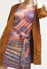 Femme portant une robe rose avec des motifs mandala colorés et une ceinture nouée à la taille, associée à une veste marron en daim avec les manches retroussées.