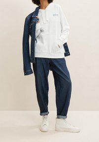 Persona que lleva una sudadera blanca con el texto "Wave", unos jeans oscuros sueltos y remangados, zapatillas blancas de caña alta, y una chaqueta de mezclilla azul oscura caída sobre un hombro.