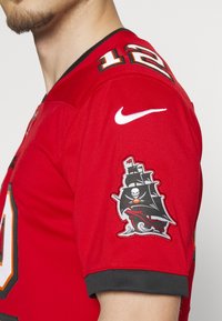 Homme portant un maillot de sport rouge avec le numéro 12 et un écusson noir avec un logo de navire pirate sur la manche et un swoosh blanc Nike au-dessus.