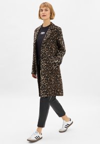 Luipaardprint lange jas met een notch-kraag, twee knopen en zijzakken, gedragen over een zwart shirt en gecombineerd met zwarte broek en sneakers.