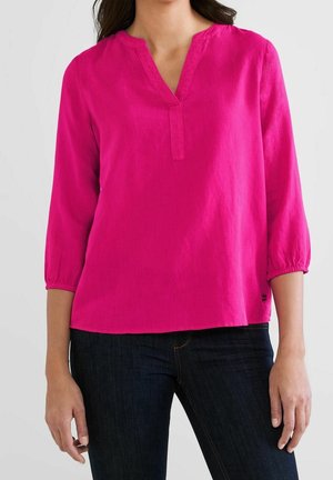Woman wearing a bright pink long-sleeve blouse with a V-neckline and dark blue jeans, standing against a light background.