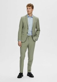 Light green tailored suit with a single-breasted blazer, matching trousers, and a light blue striped dress shirt. Black shoes complete the look.