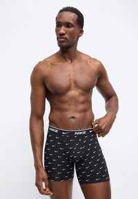 Άνδρας χωρίς πουκάμισο που ρυθμίζει μαύρα μπόξερ της Nike με λευκά λογότυπα swoosh, στέκεται μπροστά σε απλό φωτεινό φόντο.