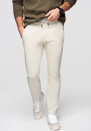 Mężczyzna w beżowych spodniach ze sznurkiem, białych trampkach i oliwkowo zielonej bluzie, z jedną ręką w kieszeni, a drugą poprawiający spodnie.
