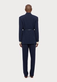 Άτομο με ξυρισμένο κεφάλι, ντυμένο με ναυτικό μπλε κοστούμι και λεπτή ζώνη, που στέκεται με την πλάτη στραμμένη προς την κάμερα σε λευκό φόντο.