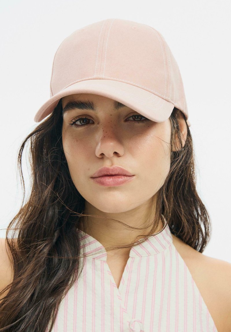Jeune femme aux longs cheveux foncés portant une casquette rose pâle et un haut rayé rose clair sans manches sur fond blanc.