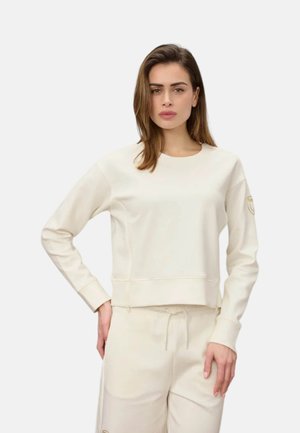 Donna con capelli castani che indossa una felpa color crema a maniche lunghe e pantaloni abbinati con coulisse, in piedi con una mano sui fianchi.
