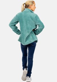 Giacca in pile turchese con colletto alto, texture morbida, vestibilità rilassata e spacchi laterali, abbinata a jeans scuri e sneakers bianche.