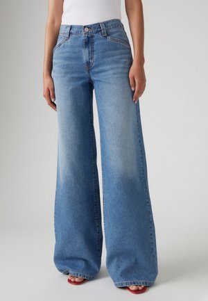 Kvinde iført højtaljede, vidde blå jeans med falmet vask og røde åbne sandaler på en ensfarvet grå baggrund.