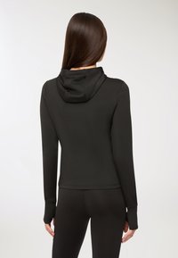 Felpa nera aderente con maniche lunghe e un cappuccio piccolo. Texture liscia, senza motivi o accessori visibili. Abbinata a leggings neri coordinati.