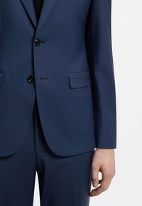Veste de costume bleu marine cintrée avec deux boutons fermés, poches à rabat, portée avec un pantalon assorti, main détendue le long du corps.