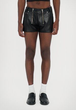 Schwarze Leder-Shorts mit Frontreißverschlüssen, taillierter Passform, flachen Taschen und einer glatten Textur. Getragen mit weißen Socken und Slippern.