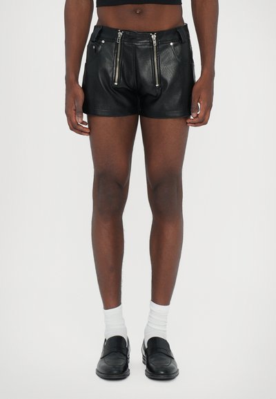 Schwarze Leder-Shorts mit Frontreißverschlüssen, taillierter Passform, flachen Taschen und einer glatten Textur. Getragen mit weißen Socken und Slippern.