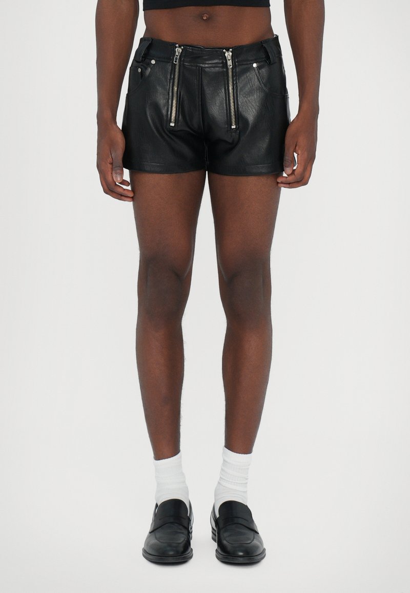 Schwarze Leder-Shorts mit Frontreißverschlüssen, taillierter Passform, flachen Taschen und einer glatten Textur. Getragen mit weißen Socken und Slippern.