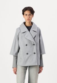 Manteau double boutonnage gris clair avec grand col, manches trois quarts, tissu texturé et deux boutons noirs. Porté par-dessus un haut noir.