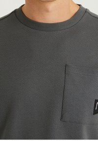 Primo piano di una persona che indossa una maglietta a girocollo grigio scuro con una texture, dotata di una tasca quadrata sul petto e un piccolo dettaglio a patch nero.