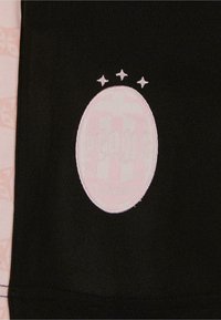 Melns audums ar ovālu rozā izšūtu logo, kuram virs tā ir trīs zvaigznes. Tekstūra ir gluda, un dizains ietver smalkus detaļas.