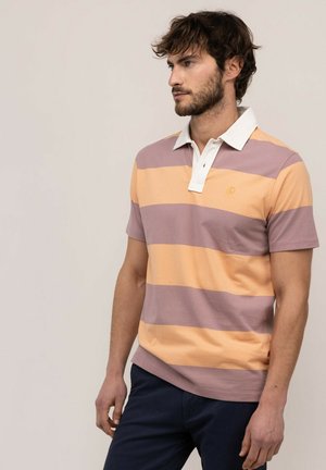 Hombre que lleva una camiseta polo de manga corta con amplias rayas horizontales naranjas y malva y pantalones oscuros, de pie frente a un fondo claro.