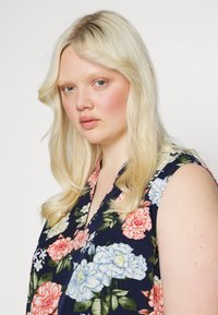 Top sem mangas florido com fundo azul-escuro, apresentando flores em tons de rosa e azul, design em forma de V e textura macia do tecido.