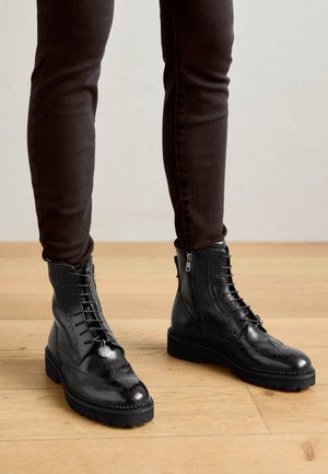 Botas de cuero negro hasta el tobillo con un diseño brogue, que presentan una cremallera frontal, cordones y una suela texturizada, combinadas con pantalones oscuros ajustados.