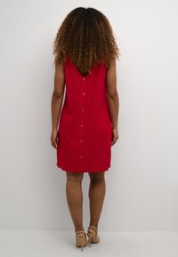 Rotes ärmelloses Kleid mit einem V-förmigen Rückendesign, das über Knopfdetails entlang des Rückens und einen lockeren Schnitt verfügt. Glatte Stoffqualität mit minimaler Textur.