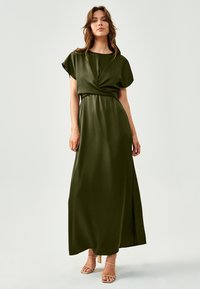Olivno zelena maxi obleka z ovito zasnovo, kratkimi rokavi in razporkom. Narejena je iz gladkega materiala in ima prilagojen pas.