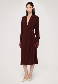 Bordowa sukienka kopertowa z głębokim dekoltem w kształcie litery V, długimi rękawami i szczegółem z klamrą po bocznej stronie, uzupełniona bordo rękawiczkami i beżowymi szpilkami.