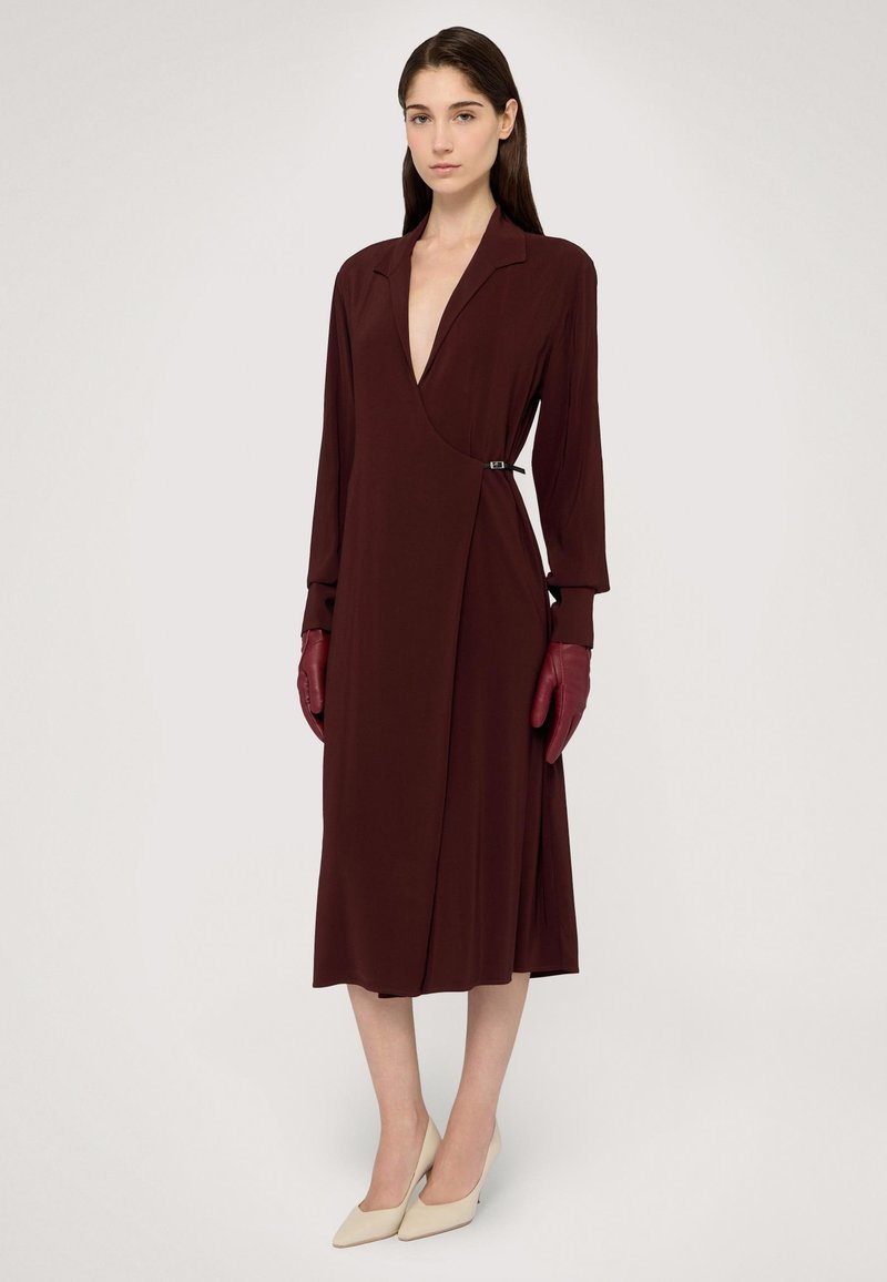 Bordowa sukienka kopertowa z głębokim dekoltem w kształcie litery V, długimi rękawami i szczegółem z klamrą po bocznej stronie, uzupełniona bordo rękawiczkami i beżowymi szpilkami.