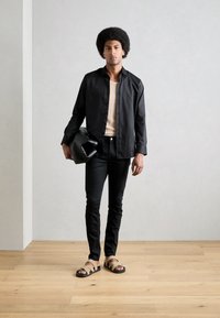 Mustat napitushirtsit, beige ribbed-top, mustat slim-fit farkut, ruskeat ja mustat sandaalit. Malli pitää mustaa, muotoiltua laukkua puulattialla.