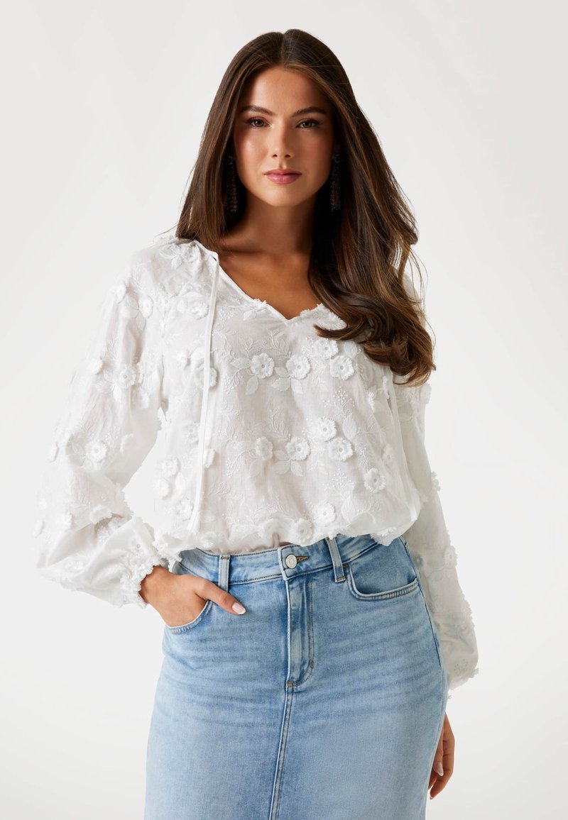 Blusa bianca ricamata con motivi floreali, scollatura con cordoncino e maniche elastiche, abbinata a una gonna di jeans blu chiaro.