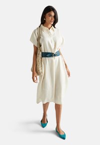 Vestido de lino color crema con mangas cortas tipo dolman, con botones al frente, ajustado con un cinturón azul verdoso. Combinado con unas bailarinas azul verdoso de punta y un bolso de mimbre.