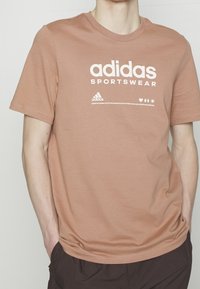 Hnědý bavlněný triko s bílým sítotiskem loga "adidas Sportswear" a malými ikonami pod ním. Krátké rukávy a kulatý výstřih.