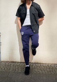 Kortärmad svart skjorta med knappar, vit linne, lila sweatpants med emblem och svarta sneakers. Outfiten har fickor och dragsko.