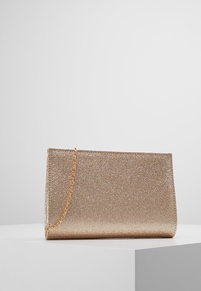 Pochette scintillante dorée de forme rectangulaire fine avec une délicate bandoulière chaîne dorée reposant sur une surface blanche.